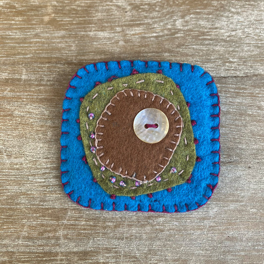 Textile handmade brooch in natural felt. Colour blue, green, brown with beads and a mother of pearl button.