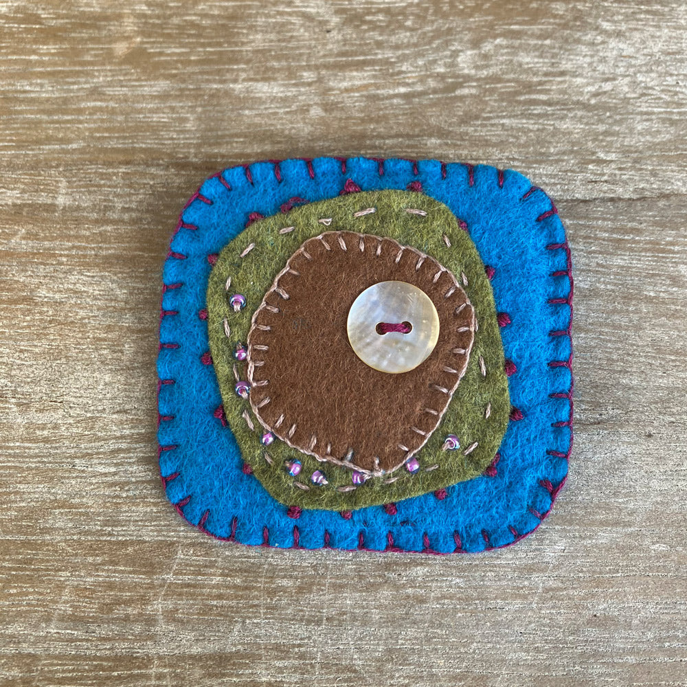 Textile handmade brooch in natural felt. Colour blue, green, brown with beads and a mother of pearl button. 